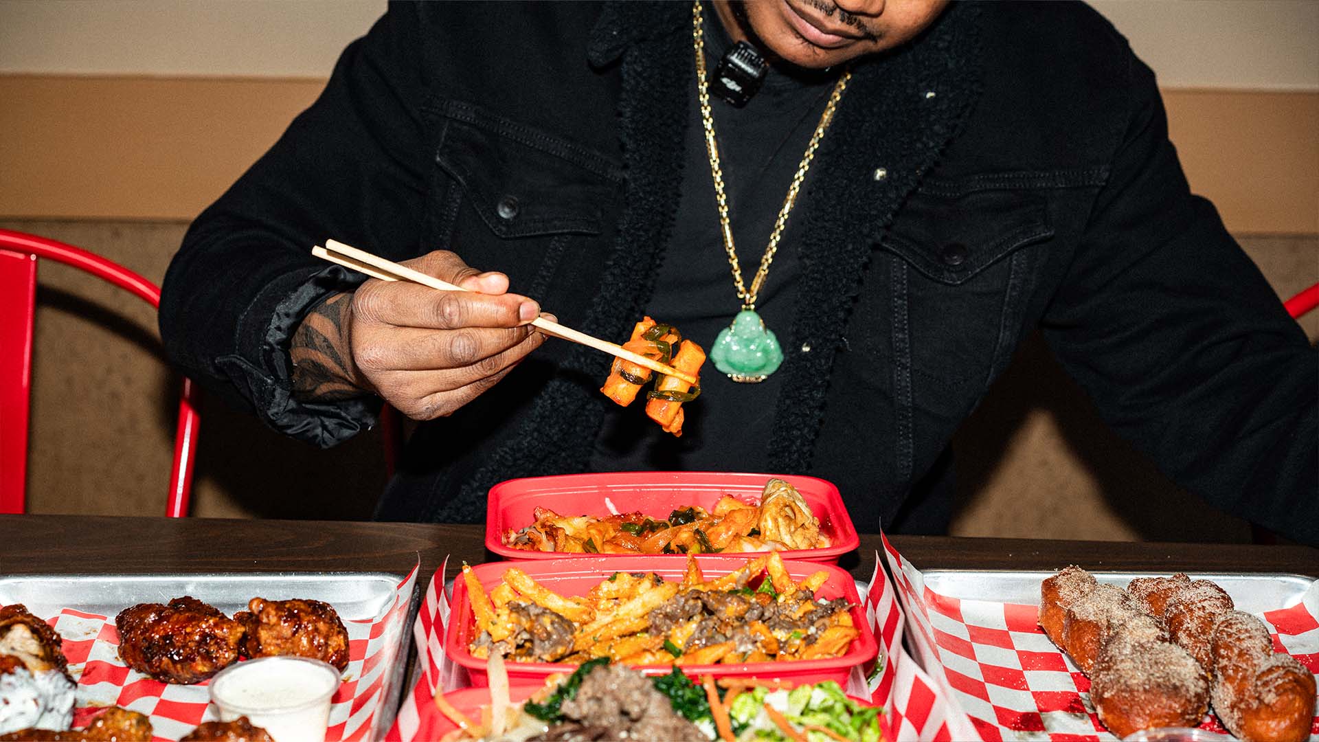 A guest picking up kimchi with chopsticks over a spread of Bonchon Chicken wings, fries, and fried treats in takeout trays.