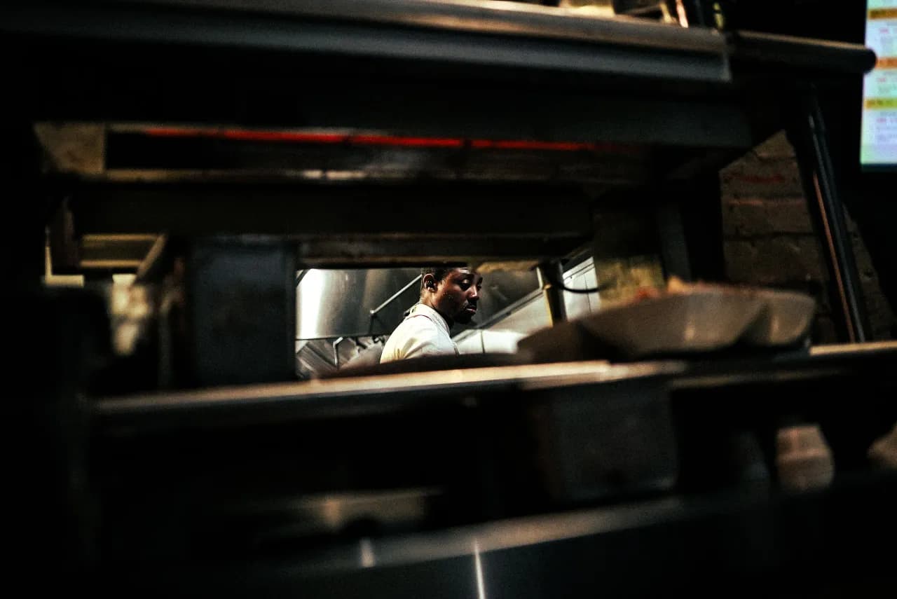 A chef in a white coat working the line, seen through the kitchen service window at Babushka's.