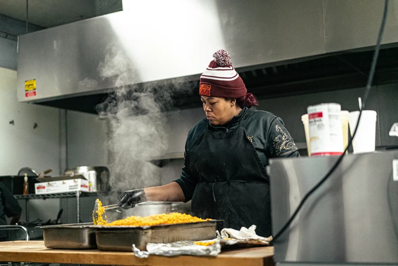 A cook in a maroon beanie plating a steaming tray of mac and cheese in the Blue Flame kitchen.