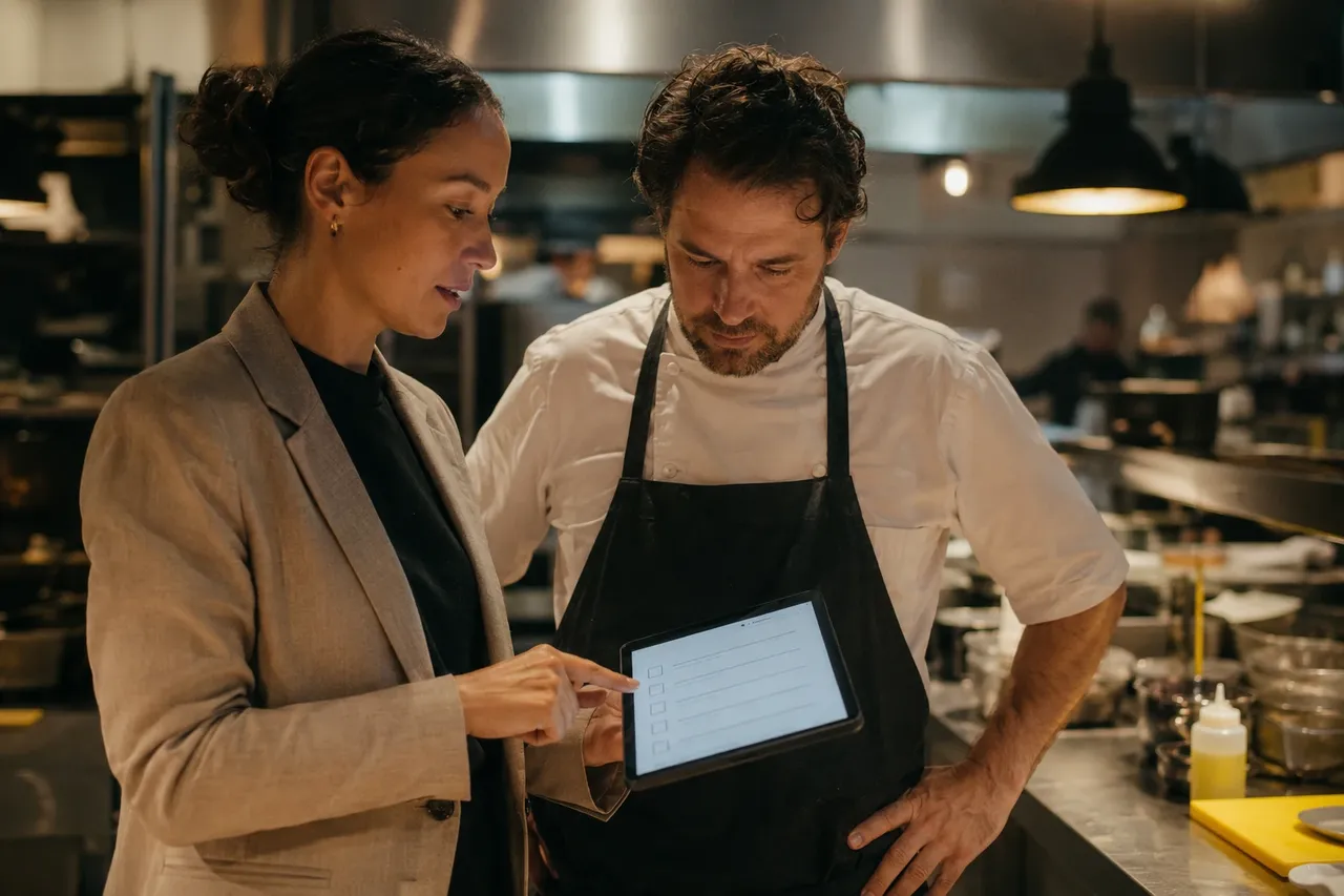 A consultant and a chef reviewing a tablet showing a recipe checklist together in a restaurant kitchen.