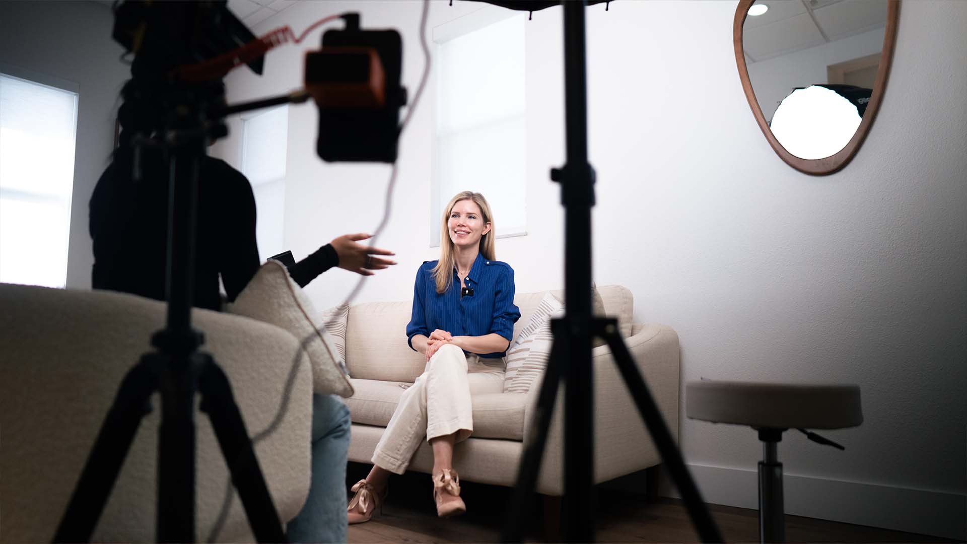 Behind the scenes of a video interview shoot: a woman seated on a couch, lit for camera, mid-conversation.
