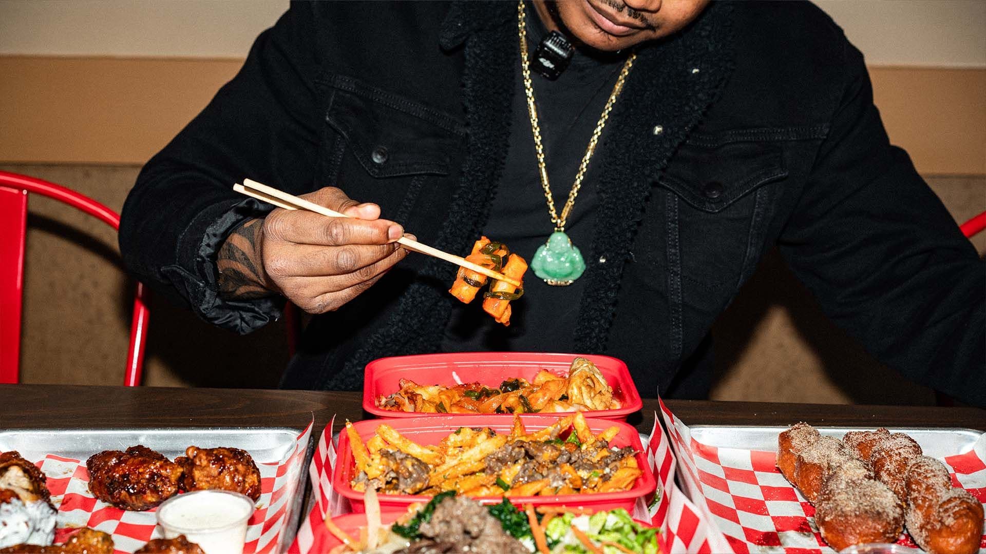 A guest picking up kimchi with chopsticks over a spread of Bonchon Chicken wings, fries, and fried treats in takeout trays.