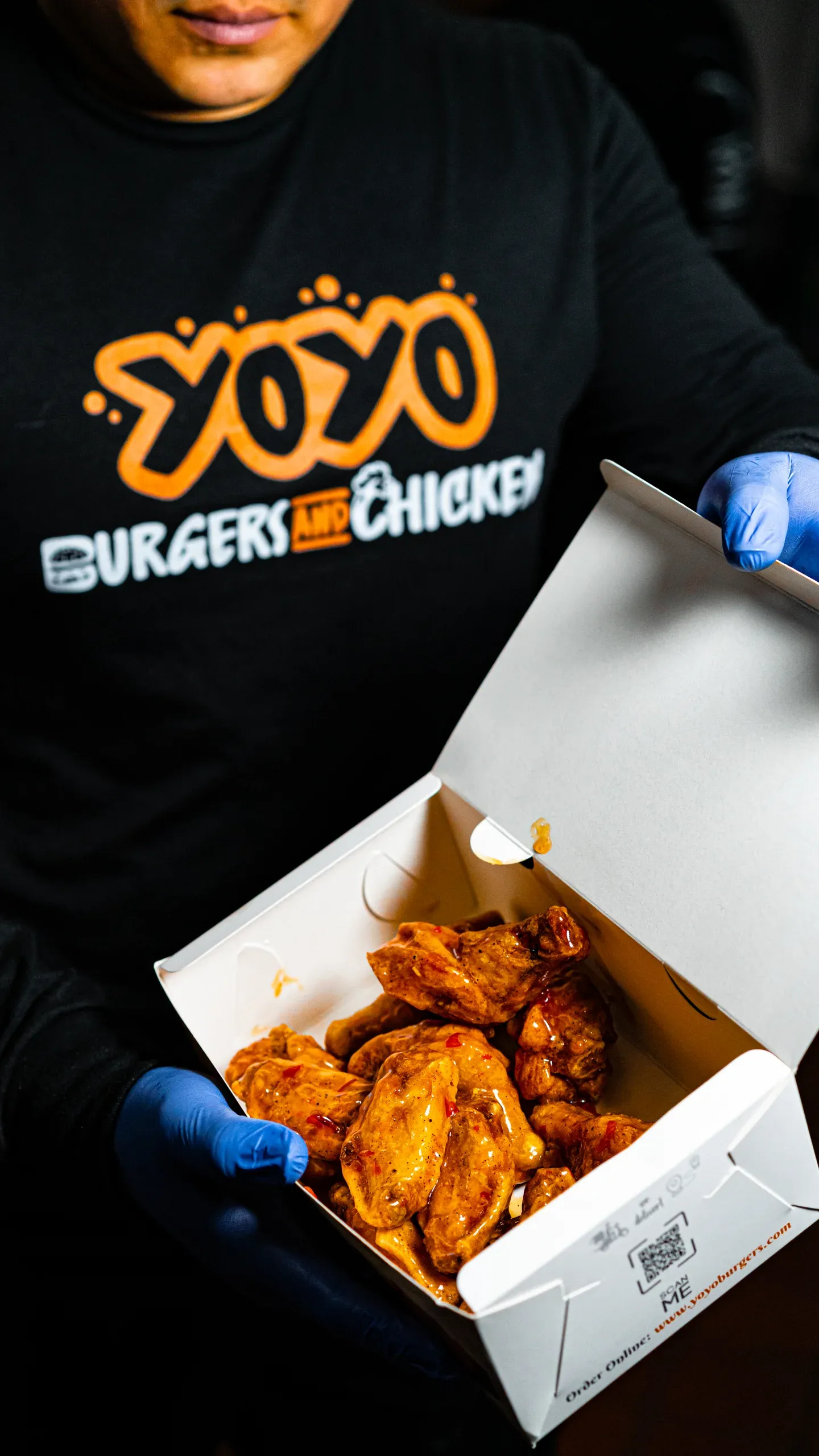 A staff member in a YoYo Burgers and Chicken hoodie holding open a takeout box of saucy wings.