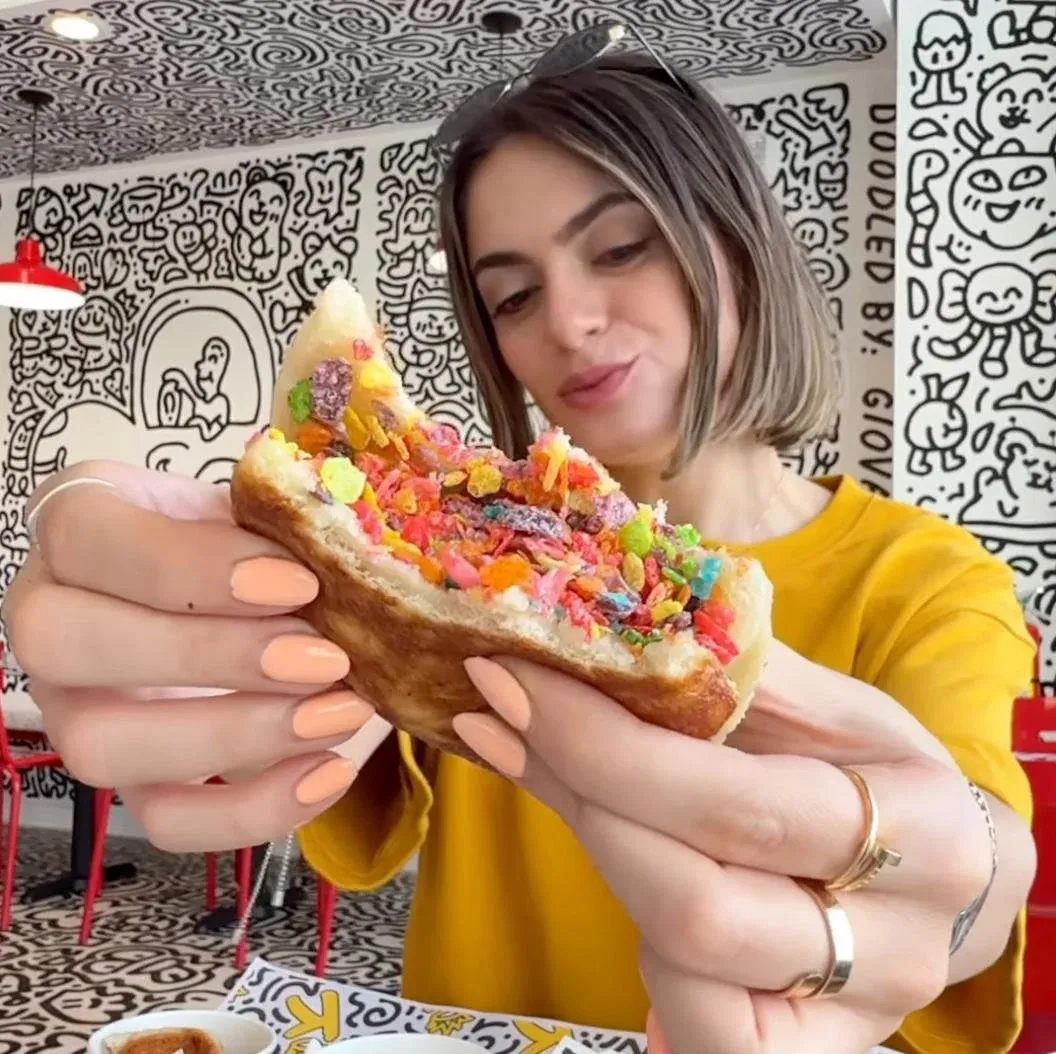 Tiana of Food by Tiana holding a cereal-crusted sandwich in a restaurant covered in doodle art