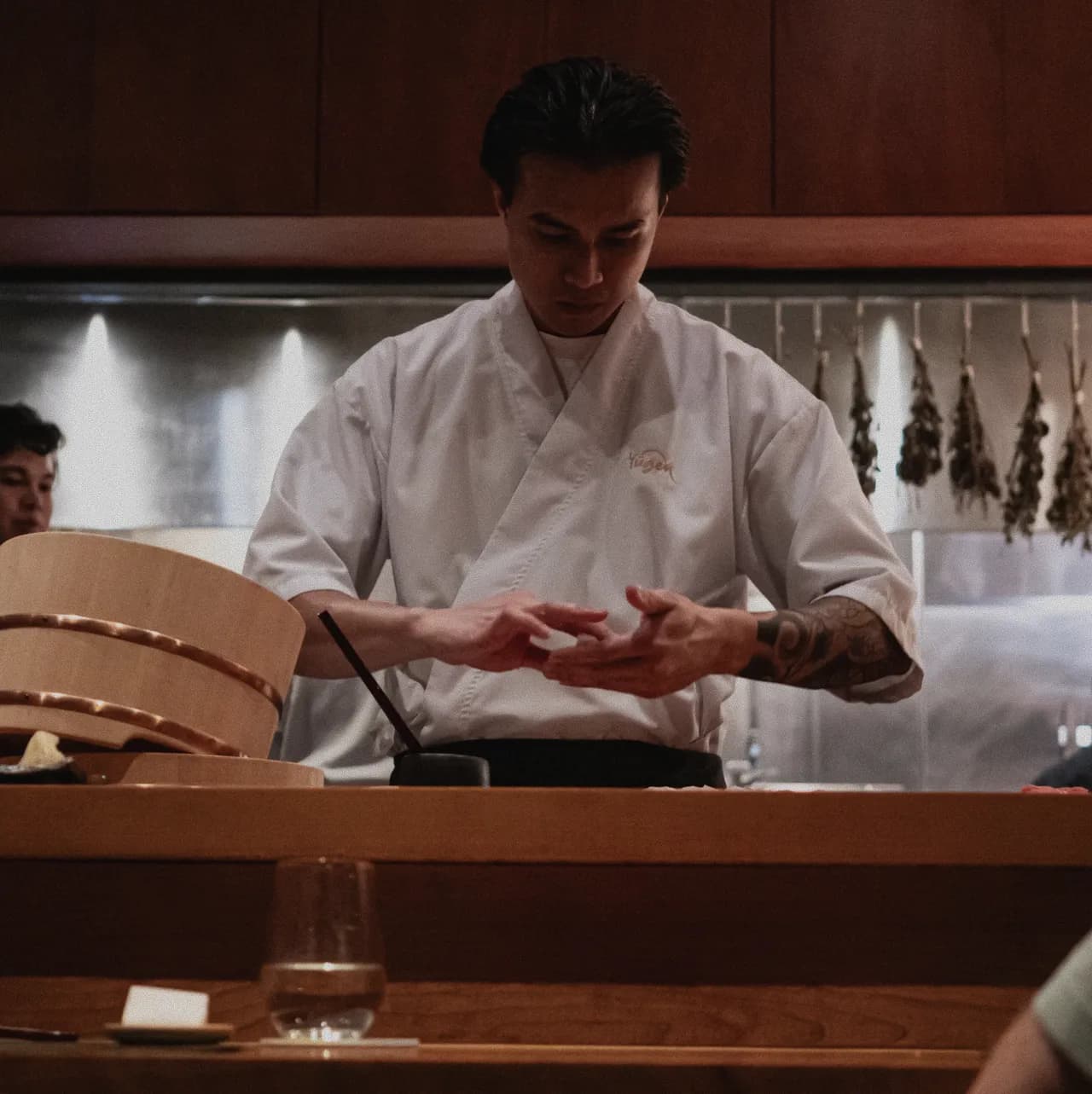 A chef in a white jacket shaping a course at the counter, the open kitchen behind