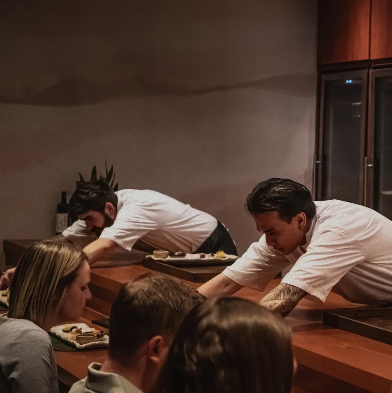 Two chefs leaning across the counter to serve guests at the Mei × Yugen dinner