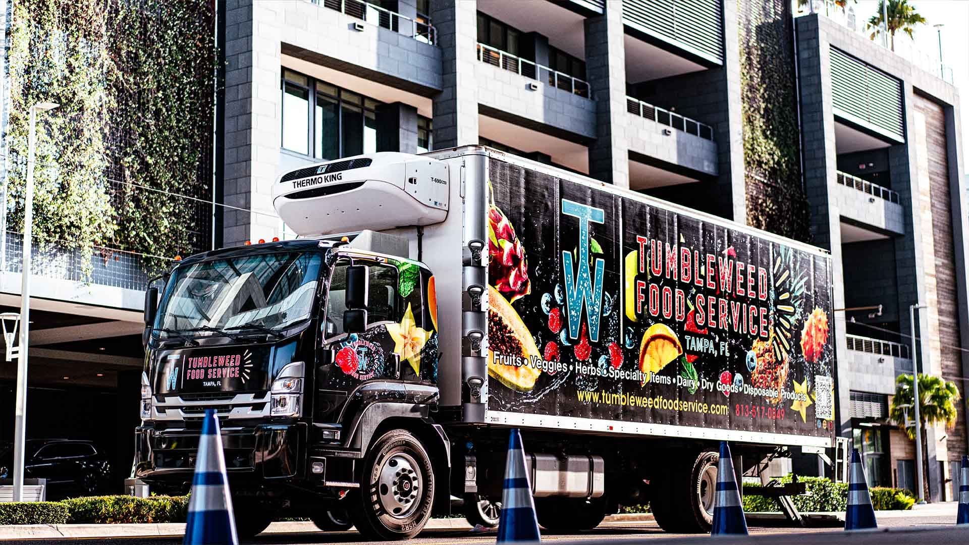 A Tumbleweed Food Service delivery truck, branded with fruit and vegetable graphics, parked outside a Tampa building.