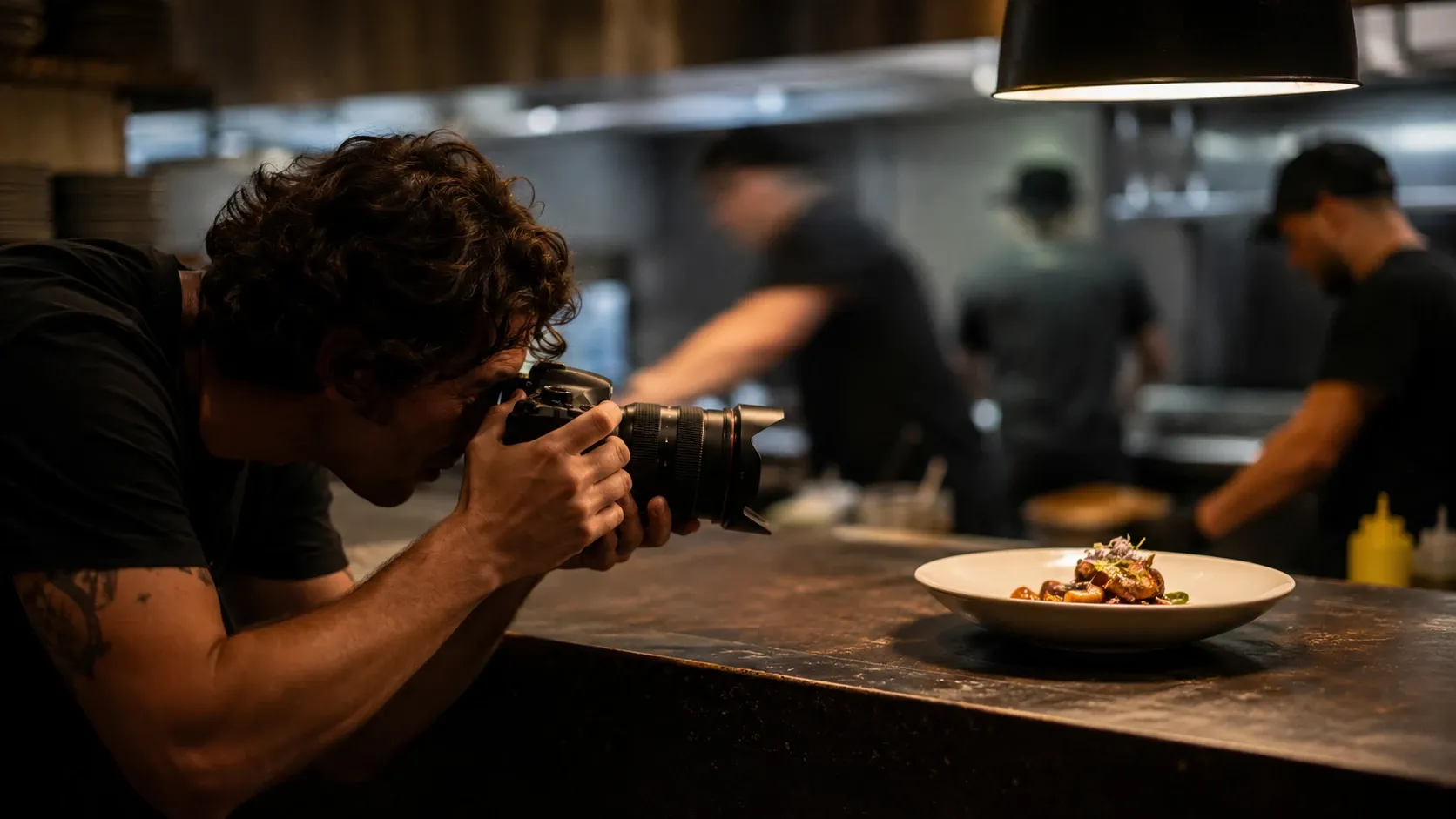 A photographer crouched low, camera in hand, photographing a plated dish on a restaurant counter mid-service.